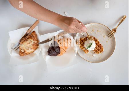 Blick von oben auf die Hand mit einem Löffel, der Eiswaffel und Croissant auf dem weißen Tisch im Café ausschaufelt Stockfoto