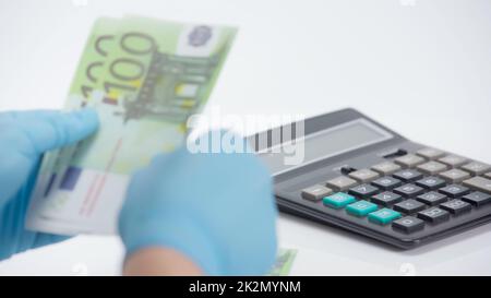 Abgeschnittene Ansicht einer Person in Latexhandschuhen, die Euro-Banknoten in der Nähe des Rechners auf weißem Hintergrund zählt Stockfoto