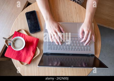 Draufsicht auf die Frau, die ihren Laptop in einem Café benutzt. Aufnahme einer jungen Frau, die mit einer Tasse Kaffee an einem Tisch sitzt und mit einem Mobiltelefon auf ihrem Laptop im Internet surft. Hochwertige Fotos Stockfoto