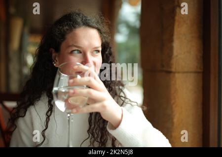 Eine glückliche Frau, die ein Glas Wasser hält, blickt durch das Fenster Stockfoto