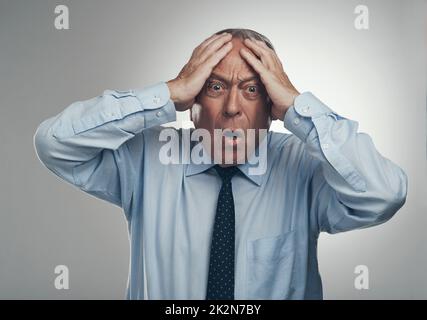 OOO, das muss weh tun. Aufnahme eines älteren Geschäftsmannes, der vor einem grauen Studiohintergrund mit dem Kopf in den Händen steht und schockiert aussieht. Stockfoto