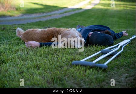 Junger Mann und sein Hund liegen im Gras mit Krücken im Vordergrund Stockfoto