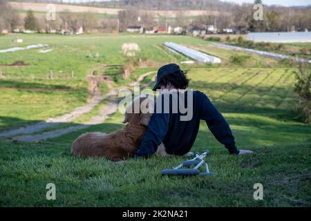 Der junge Mann kuschelt den alten Golden Retriever auf einem Hügel mit Krücken auf dem Boden Stockfoto