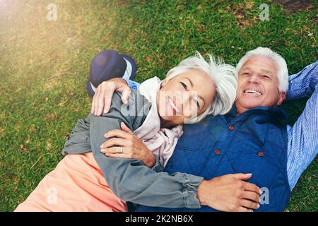 Jeden Tag verliebe ich mich wieder in ihn. Porträt eines glücklichen älteren Paares, das sich im Park auf dem Gras niederlegt. Stockfoto