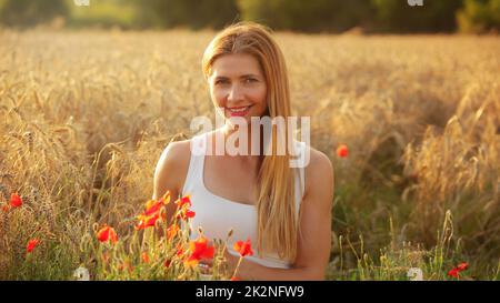 Junge Frau sitzt im Weizenfeld, beleuchtet durch die Nachmittagssonne, Paar roter Mohn um Sie im Vordergrund. Stockfoto