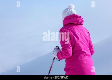 Von hinten betrachten, junge Frau in rosa Winterjacke, Stöcke in die Hände, um den Hügel hinunter zu gehen. Stockfoto