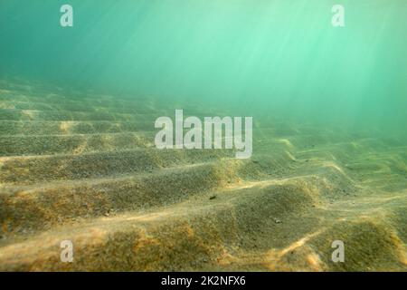 Unterwasserfoto, kleine Sanddünen schossen diagonal, so dass sie aus dieser Perspektive Treppen bilden, Sonnenstrahlen kommen von der Meeresoberfläche. Abstrakter Meereshintergrund. Stockfoto