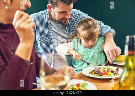 Ich kann es selbst machen, Dardist. Aufnahme eines fröhlichen jungen Mannes, der seinen Töchtern beim Essen half, während er zu Hause an einem Esstisch saß. Stockfoto