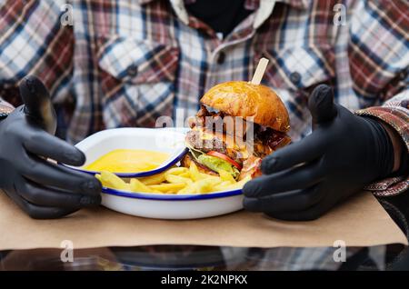 Das Konzept von Fast Food und Essen zum Mitnehmen. Ein Mann in schwarzen Latexhandschuhen ist bereit, einen saftigen Hamburger zu essen, der neben den pommes frites auf einem Metallteller liegt, zusammen mit Käsesauce. Stockfoto