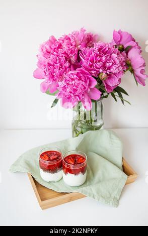 Ein schönes Bouquet aus rosa Pfingstrosen in einer Vase auf einem weißen Tisch steht zusammen mit einem herzförmigen Erdbeerdessert. Valentinstag, 8. März. Stockfoto