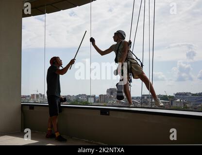 Professioneller Bergsteiger-Reiniger, der am Seil hängt und das Fenster mit einem Lappen abwischt, während ein Kollege im Gebäude steht und auf das schmutzige Fenster zeigt. Stockfoto