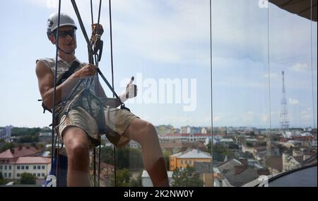 Industriebergsteiger Arbeiter in Schutzhelm zeigt Zustimmung Geste und lächeln, während sie an Seil hinter Fenster hängen. Mann, der Sicherheitshebegeräte außerhalb des Gebäudes verwendet. Stockfoto