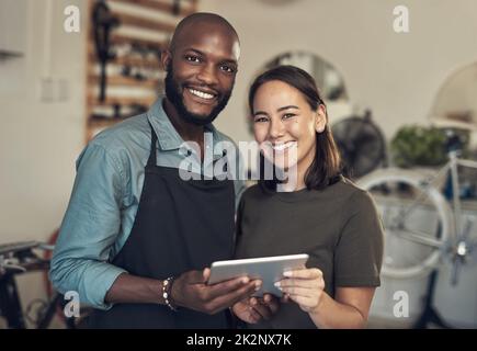 Lassen Sie uns Ihnen bei der Auswahl Ihres Fahrrads helfen. Aufnahme von zwei jungen Geschäftsbesitzern, die in ihrem Fahrradladen zusammenstehen und ein digitales Tablet benutzen. Stockfoto