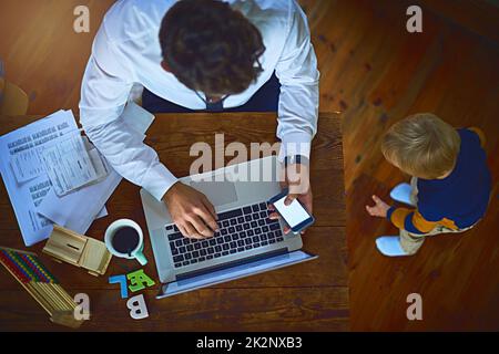 Er arbeitet, behält aber immer noch sein Baby im Auge. Foto eines unbekannten alleinerziehenden Vaters, der sein Handy und seinen Laptop benutzte, während er seinen Sohn zuhause beobachtete. Stockfoto