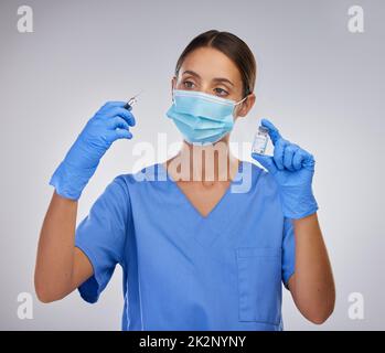 Schützen Sie andere durch Impfungen. Aufnahme einer jungen Krankenschwester, die eine Nadel und eine Ampulle mit Impfflüssigkeit vor einem Studioraum hält. Stockfoto