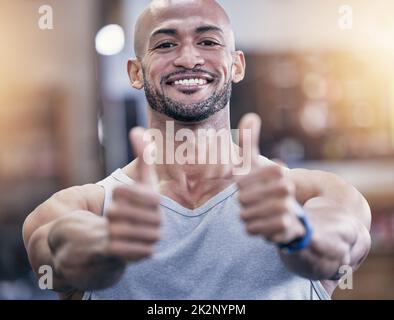 Hier unterstützen Sie Ihre lebensverändernde Fitness-Reise. Porträt eines muskulösen jungen Mannes, der in einer Turnhalle die Daumen hochzeigt. Stockfoto