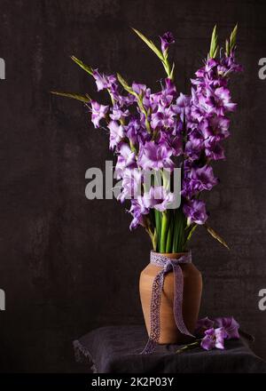 Stillleben mit schönen Bouquet von lila, lila Gladioli Blumen in Keramikvase. (Gladiolus) Dunkler rustikaler Stil. Heim- oder Gartendekoration. Stockfoto