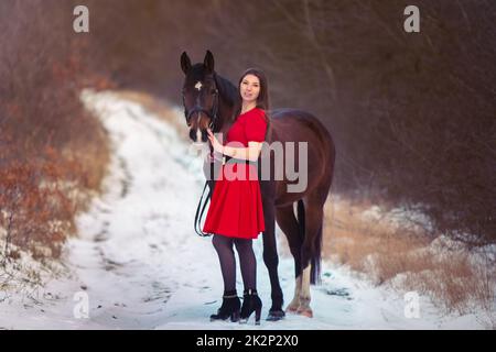 Ein schönes Mädchen in einem roten Kleid geht mit einem Pferd vor der Kulisse eines Winterwaldes Stockfoto