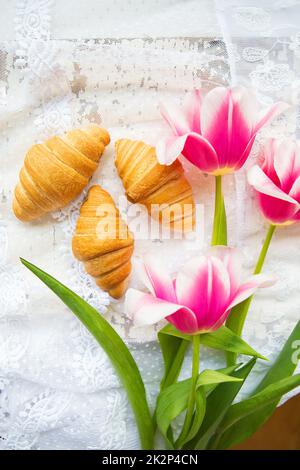 Drei Croissants und leuchtend rosa Tulpen auf Spitze Tischdecke Stockfoto