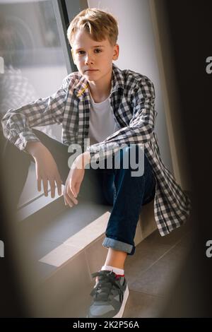 Ein stilvoller Teenager in legerer Kleidung blickt auf die Kamera, während er in einem Gebäude in der Nähe des Fensters auf dem Boden sitzt Stockfoto