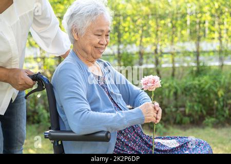 Altenpfleger Tochter umarmen und helfen asiatische ältere oder ältere alte Dame Frau mit roten Rose auf Rollstuhl im Park. Stockfoto