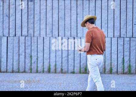 Stilvoller Mann mit Hut und Mobiltelefon, während er draußen auf der Straße unterwegs ist. Technologiekonzept. Stockfoto