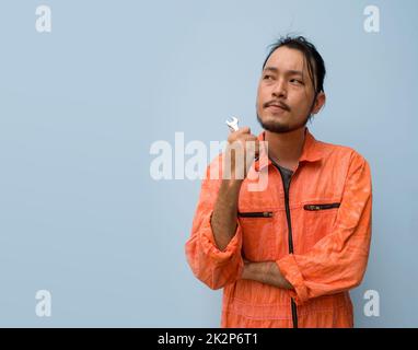 Der Chefmechaniker in einer orangefarbenen Uniform hielt einen kleinen Schraubenschlüssel. Er stand mit gekreuzten Armen auf dem blauen Bildschirm. Porträt mit Studiolicht. Stockfoto