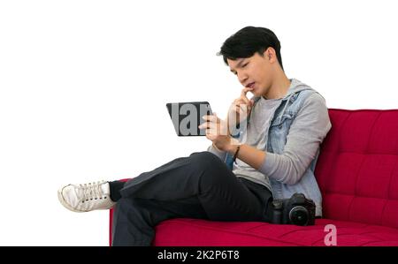 Der junge asiatische Fotograf sieht sich Bilder an, die auf dem Tablet-Computer-Bildschirm aufgenommen wurden, während er auf einem roten Sofa sitzt. Arbeitsatmosphäre im Fotostudio Stockfoto