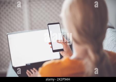 Frau mit Smartphone in den Händen sitzt drinnen und tippt auf dem Laptop Stockfoto
