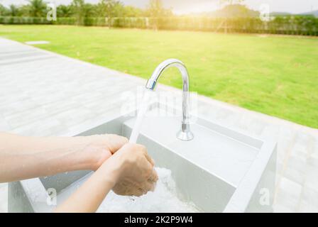 Frau wäscht sich die Hände mit Leitungswasser unter Wasserhahn am weißen Waschbecken. Waschen Sie die Hände mit Leitungswasser am Waschbecken im Freien in der Nähe des Grasfeldes. Persönliche Hygiene zur Vorbeugung von Coronavirus oder Covid 19. Gesunder Lebensstil. Stockfoto