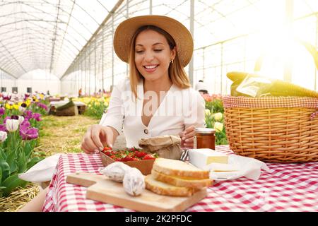 Lächelnd schönes Mädchen mit Picknick. Junge Frau nimmt eine Erdbeere. Frische Ernte und Null-Kilometer-Lebensmittel im Land im Frühling. Stockfoto