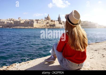 Reisekonzept mit unabhängigen Personen, die die Outdoor-Aktivitäten und den Lebensstil des Wanderessens genießen. Eine junge Frau sitzt am Meeresrand und sieht die atemberaubende Landschaft von Valletta, Malta, aus.
