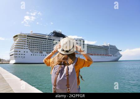 Junge Backpacker-Frau mit Strohhut, die vor einem großen Kreuzfahrtschiff steht Stockfoto