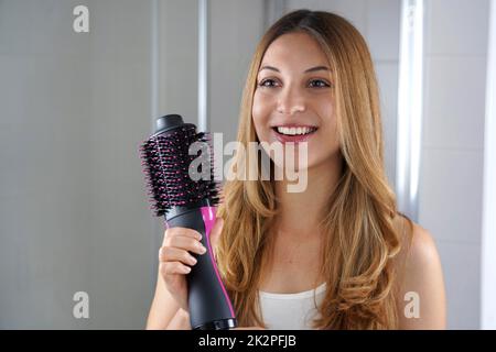 Das wunderschöne Mädchen hält einen runden Pinsel-Haartrockner, um die Haare zu Hause zu stylen. Junge Frau zeigt einen einstufigen Haartrockner und Volumenizer im Salon. Stockfoto