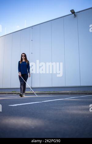 Junge Frau mit Sehbehinderung geht auf den Straßen der Stadt und nutzt ihren weißen Stock, um besser durch den städtischen Raum zu navigieren und sicher an ihr Ziel zu gelangen Stockfoto