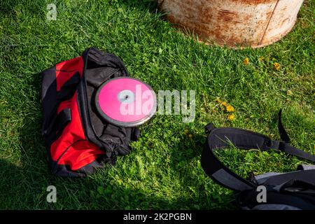 Rote Sporttasche auf dem Rasen mit pinkfarbenem Schienen- und Felddiskus Stockfoto