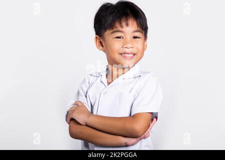 Asiatisches Kleinkind lächelt fröhlich und trägt Thai-Uniform, rote Hosen stehen mit zusammengeklappten Armen Stockfoto