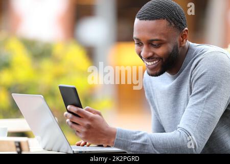 Ein Mann mit schwarzer Haut, der Handy und Laptop in einer Bar überprüft Stockfoto