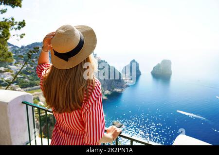 Wunderschöne junge Modefrau mit gestreiftem Kleid und Hut auf Capri Island mit Faraglioni-Meer und blauem kristallklarem Wasser im Hintergrund, Capri, Italien Stockfoto