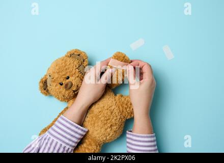 Weibliche Hand hält einen braunen Teddybär und klebt einen medizinischen Klebstoff-Gips auf blauem Hintergrund, Tram-Behandlung Stockfoto