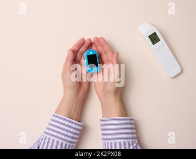 Weibliche Hand hält berührungsloses Thermometer und Oximeter aus Kunststoff auf beigem Hintergrund Stockfoto