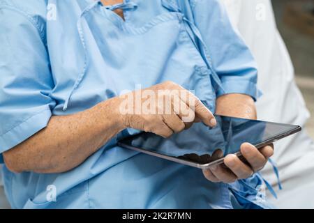 Asiatische Seniorin oder ältere Frau Patientin Patient hält in ihren Händen Digitaltablett und liest E-Mails während sie auf der Pflegestation im Bett sitzt, gesund starke medizinische Konzept Stockfoto