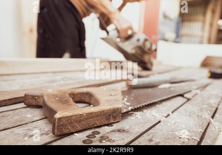 Handsäge mit Holzgriff auf verschwommenem Tischler, der mit elektrischer Hobelmaschine arbeitet. Zimmermannswerkzeuge. Handsäge und Sägemehl auf Holztisch in der Außenwerkstatt. Ein Handwerker, der Holzarbeiten macht. Handwerkkonzept. Stockfoto