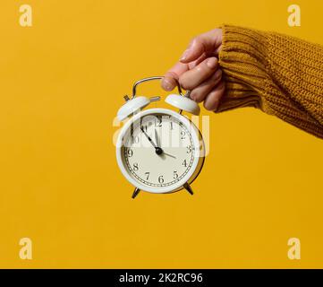 Eine weibliche Hand hält einen Wecker aus weißem Metall auf einem weißen Tisch. Konzept des Termins für den Auftrag Stockfoto