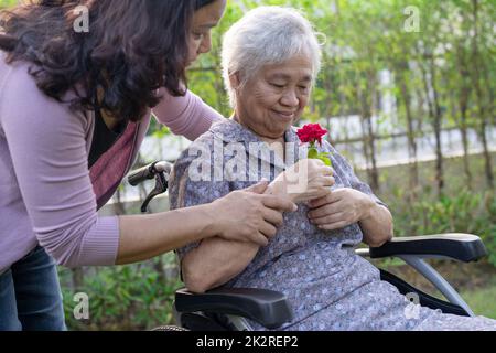 Altenpfleger Tochter umarmen und helfen asiatische ältere oder ältere alte Dame Frau mit roten Rose auf Rollstuhl im Park. Stockfoto