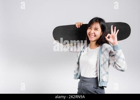 Lächelnde glückliche Frau, die das Skateboard auf der Schulter hält und das OK-Zeichen zeigt