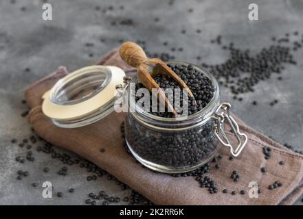 Glasbehälter aus trockenen schwarzen Linsen Bohnen mit einer Schaufel auf grauem Tisch Nahaufnahme, gesunde Protein-Diät Stockfoto