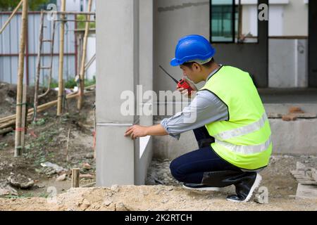 à¹à¹Young asiatischer Mann mit Schutzhelm und Sicherheitsweste, der die Hausstruktur überprüft, während er den Walkie-Talkie hält. Arbeitsumfeld von Ingenieuren auf der Baustelle von Wohnungsbauprojekten Stockfoto