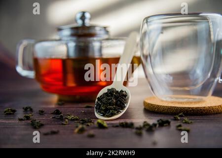 Ein Holzlöffel mit Tee liegt vor einem dunklen Holzhintergrund, ein Becher und eine Teekanne sind von hinten sichtbar, ein Foto in dunklen Farben. Stockfoto