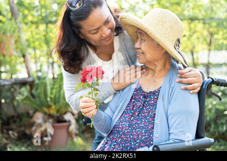 Altenpfleger Tochter umarmen und helfen asiatische ältere oder ältere alte Dame Frau mit roten Rose auf Rollstuhl im Park. Stockfoto